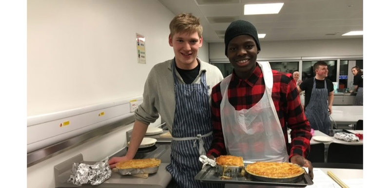 Two male students pose for a photo during cooking class, the food they cooked sits on trays and in containers in front of them.
