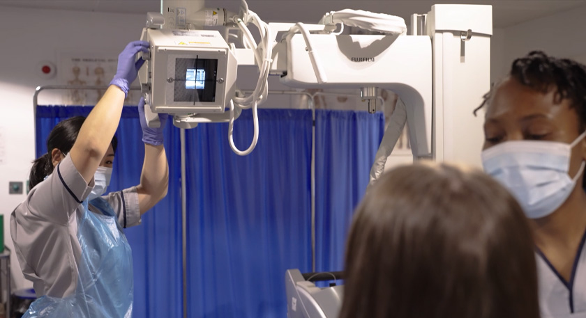 A female student operating a piece of diagnostic radiography equipment while another female student stands in front of it.
