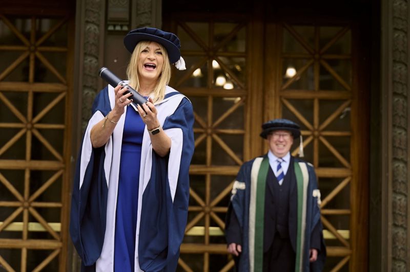 Sally Gray in her graduation gown and Paul Grice stood in front of a glass framed wooden door