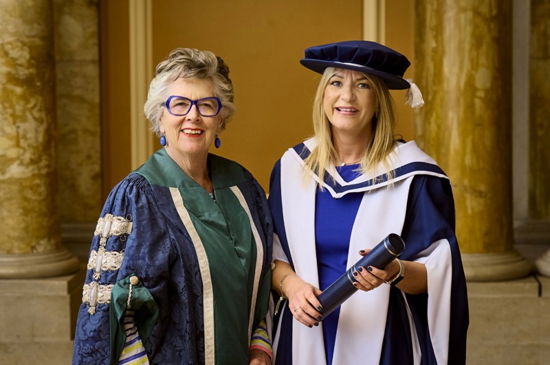 Sally Gray in her graduation gown and Paul Grice stood in front of a glass framed wooden door