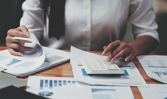 A woman using a calculator with financial reports surrounding her on a desk. 