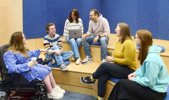 A group of six students are sat on the tiered seating in the library presentation space, having a laugh with one another.