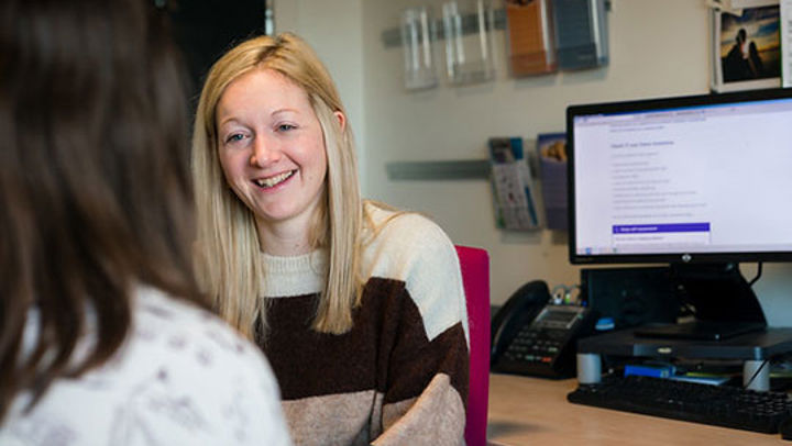 Two women talking by a computer desk, the woman in view is smiling kindly at the other