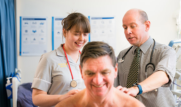 A student nurse with a doctor examining a patient's back while they all smile