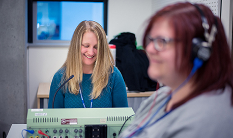 A woman with a headset on and a women behind her sat at a desk