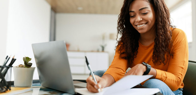 A student sitting at a low-profile desk, writing notes in front of a laptop.