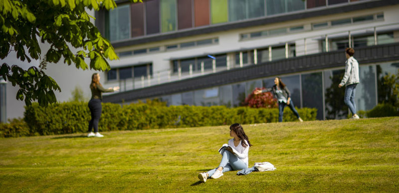 A wide angle shot of a young woman sitting on the grass beside the main QMU academic building