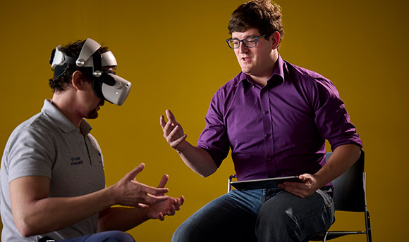 A man is seated, demonstrating the movement required in a VR paramedic simulation. They are sat alongside another man who is wearing a VR headset.