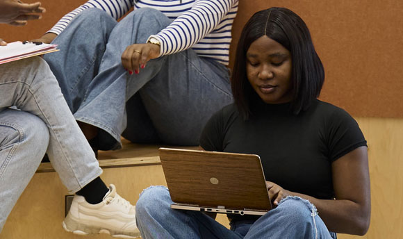 A female student using a laptop, two other students sit on the raised seating behind them.