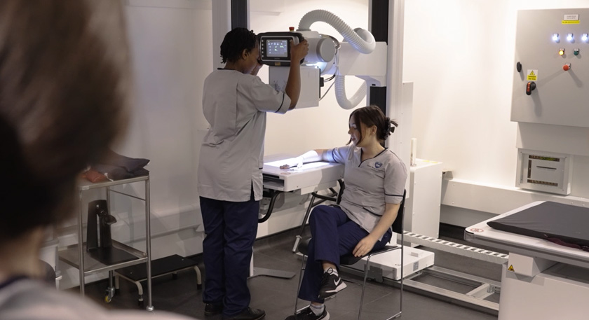 A female student operating a piece of diagnostic radiography equipment while another female student sits with their hand beneath the scanner.
