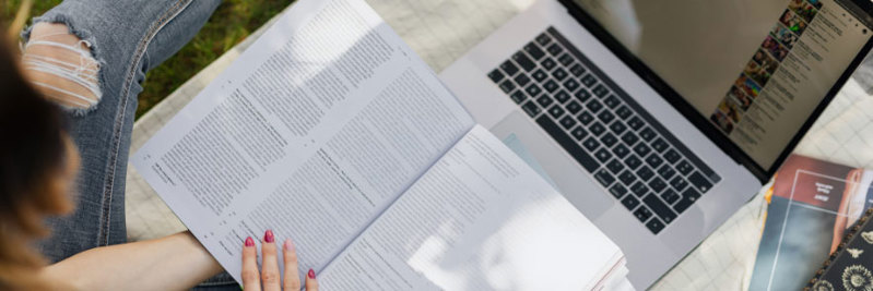 A close up of a student using a laptop while holding open a textbook on their lap.