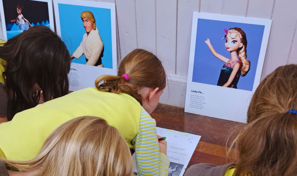 A group of children drawing on pieces of paper, some images of popular dolls and toys with visible limb differences are on display.