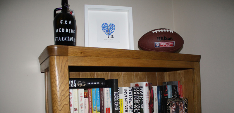A bookshelf filled with books and topped with an American football and a flask labelled "G&A Wedding Fund"