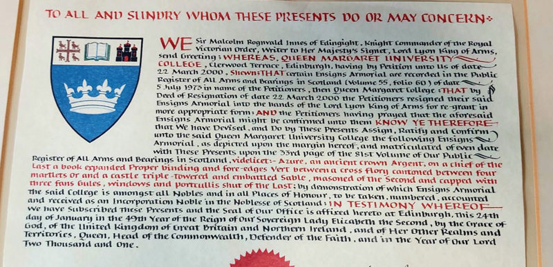The Queen Margaret University Blazon, which reads: "Azure, an ancient crown Argent, on the chief of the last a book expanded proper binding and fore edges vert between a cross flory cantoned between four martlets or and a castle triple-towered and embattled sable, masoned of the second and capped with three fans gules, windows and portcullis shut of the last."