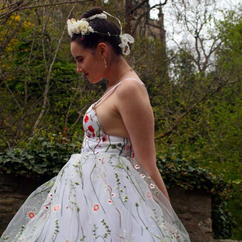A young woman wears a white strappy dress with a tulle skirt. The fabric is embroidered with colourful flowers.