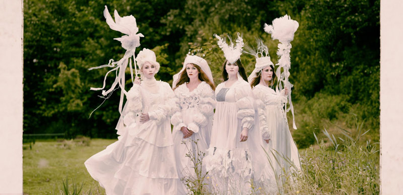 Four women wearing ornate, all-white robes, feathered hairpieces and holding white staffs. Their outfits are themed around 'Selene, goddess of the moon'.
