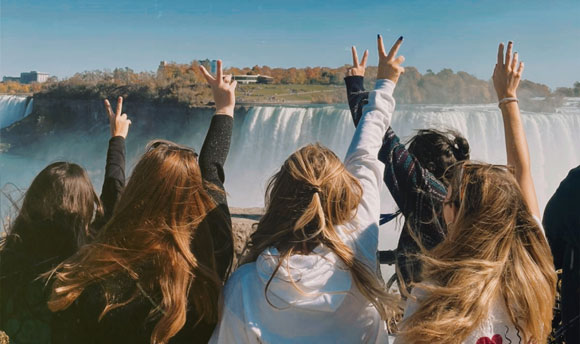 A group of young women face towards Niagara Falls (away from the camera) and hold peace signs in the air.