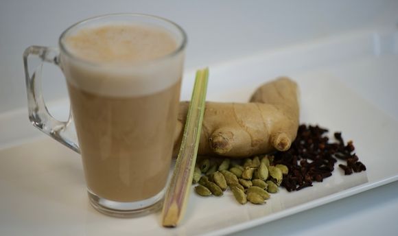 Chai tea in a clear coffee glass with cut lemongrass, ginger, cardamom and tea leaves beside