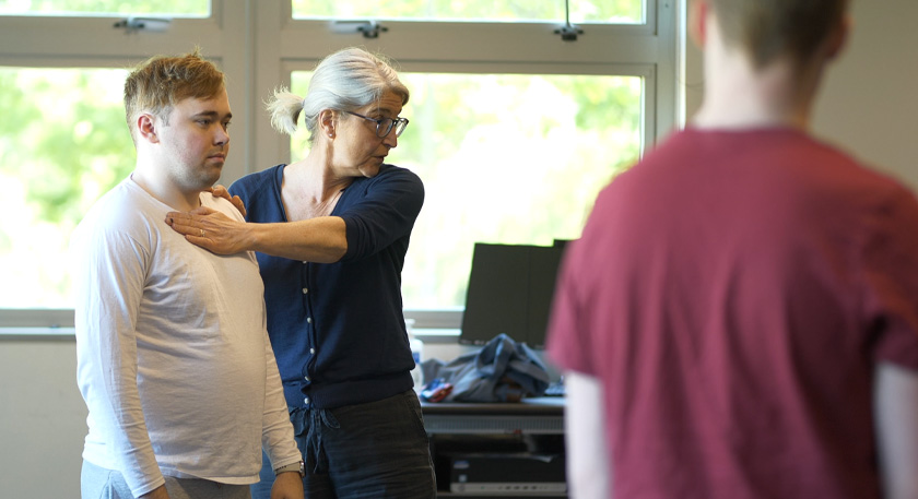 A teacher demonstrates a breathing exercise with their arm in front of a student's shoulders.