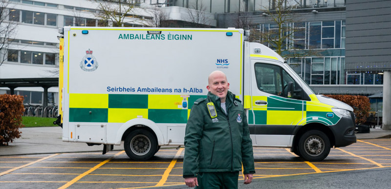 A man wearing a paramedic uniform is seen standing in front of an ambulance in front of the QMU campus entrance.