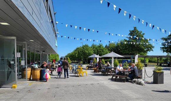 People sitting at picnic tables on a sunny day with blue skies outside Queen Margaret University's Students' Union building