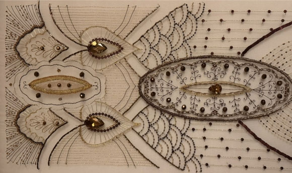 A close up of a hand-embroidered piece of fabric. The item is cream in colour, while the beads are a variety of shades including gold, brown, and black.