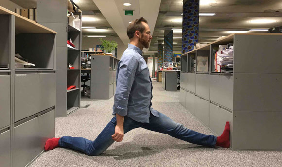 Anthony Rooke performing the splits between two shelving units in an office area