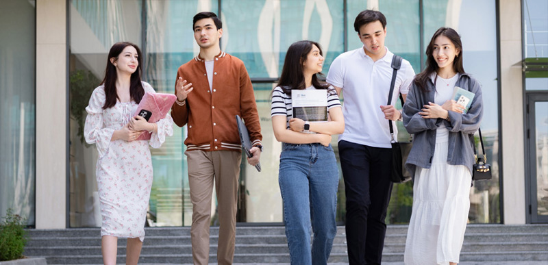 A group of two male and three female students walking out of the BMU campus. They are holding an assortment of stationery and chatting to one another as they walk.