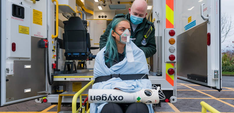 A person sits in a wheelchair, wearing a blanket and oxygen mask, while being tended to by a paramedic. Behind them is an ambulance with the doors wide open.
