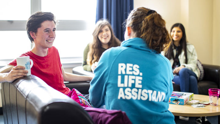 Three students sitting in the student accommodation. Facing away from the camera is a fourth student wearing a hooded sweatshirt that says "Res. Life Assistant".