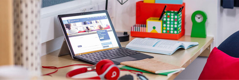 A laptop on a student's desk, the screen is open on the QMU website.