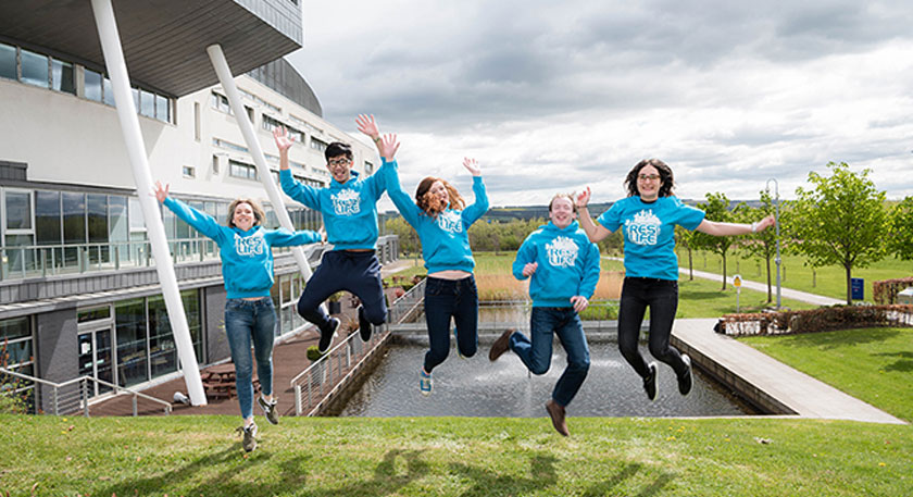 A group of students in matching blue sweaters jumping for a photo outside QMU