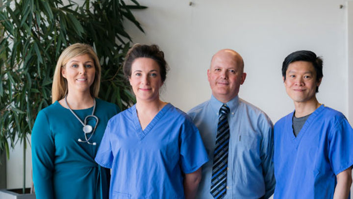 Four medical practitioners wearing blue scrubs stand smiling at the camera against a plain wall, with a large plant in the background.