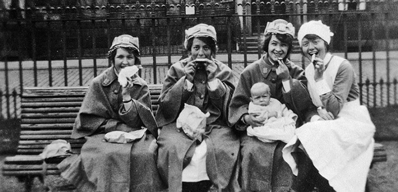 A black and white photo of four women on a bench eating lunch and smiling at the camera.