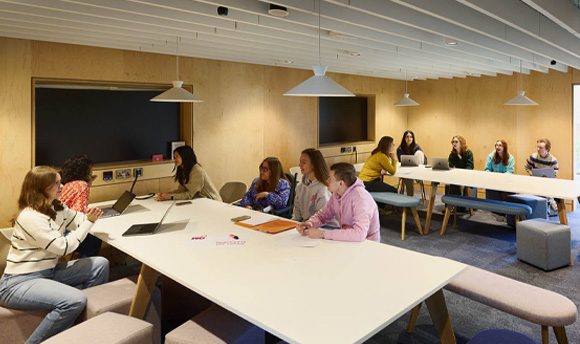 A teaching space within the library, there are two large tv screens, a whiteboard, and two long tables filled with students working on laptops.