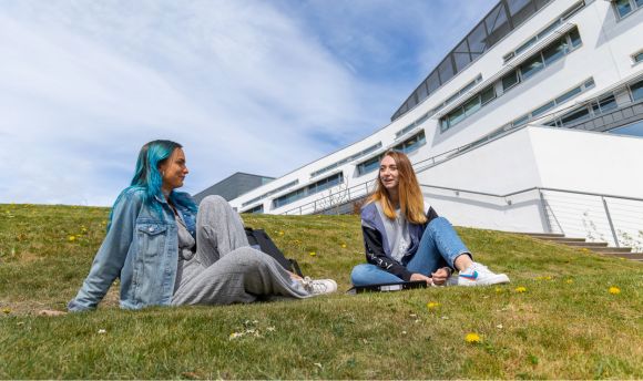 Two young women sit on the grass outside the main QMU building