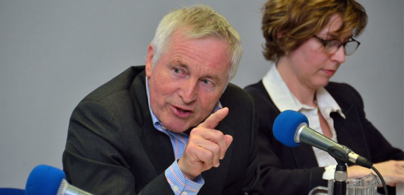 A man and a woman sat at a debate panel, the man is pointing towards the microphone in front of him.