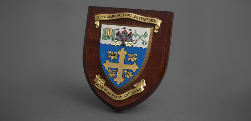 The Queen Margaret University College crest and motto, "with Head, Heart and Hand" affixed to a wooden shield.