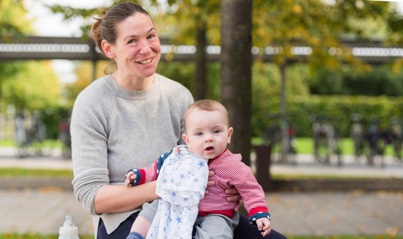 A woman holding an infant on her lap sits on a bench in QMU square
