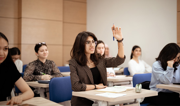 A young woman holds her hand up to ask a question, seated at an individual work desk in a classroom surrounded by other students.