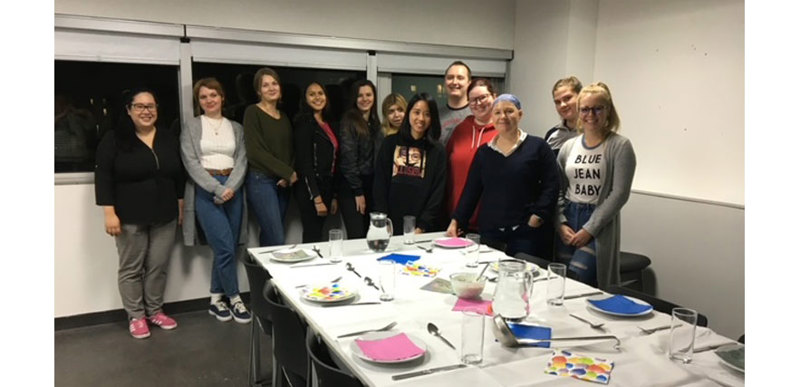 A group of students pose for a photo behind a table laid out with dinnerware and cutlery for a sit-down meal.