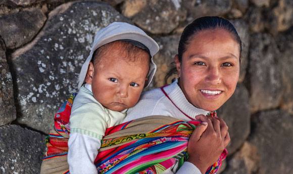A young woman carries an infant on her back, tied securely with a brightly coloured fabric.