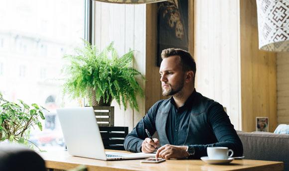 A male student working at laptop beside large bright window with plants.