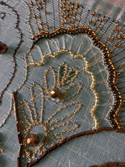 A close up of a hand-embroidered piece of pale blue fabric. The beads are gold and brown in colour.